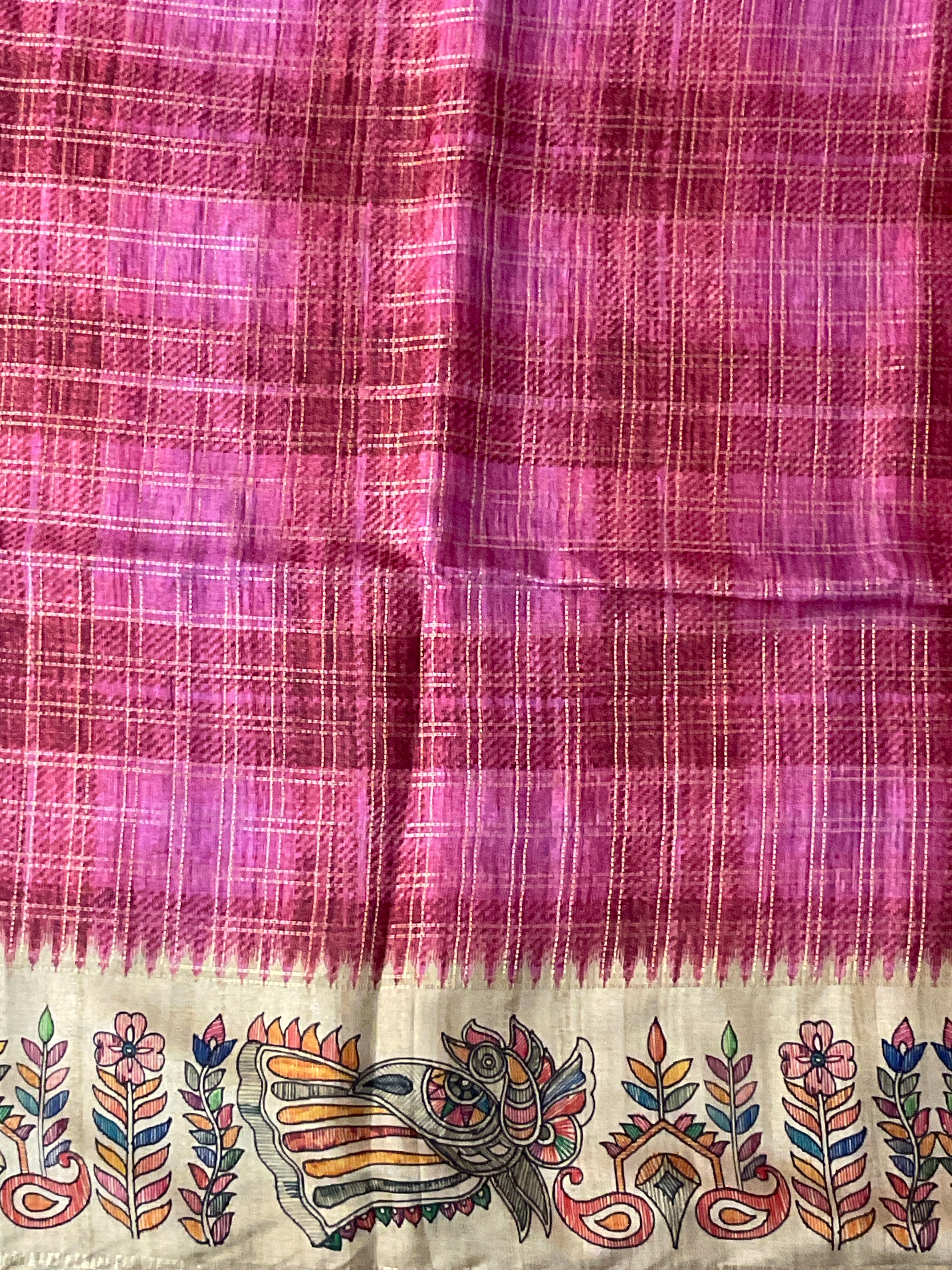 A close-up of a pink and magenta plaid fabric, a saree, featuring a decorative Kalamkari border with colorful hand-painted motifs including a central bird and floral elements on a cream background.