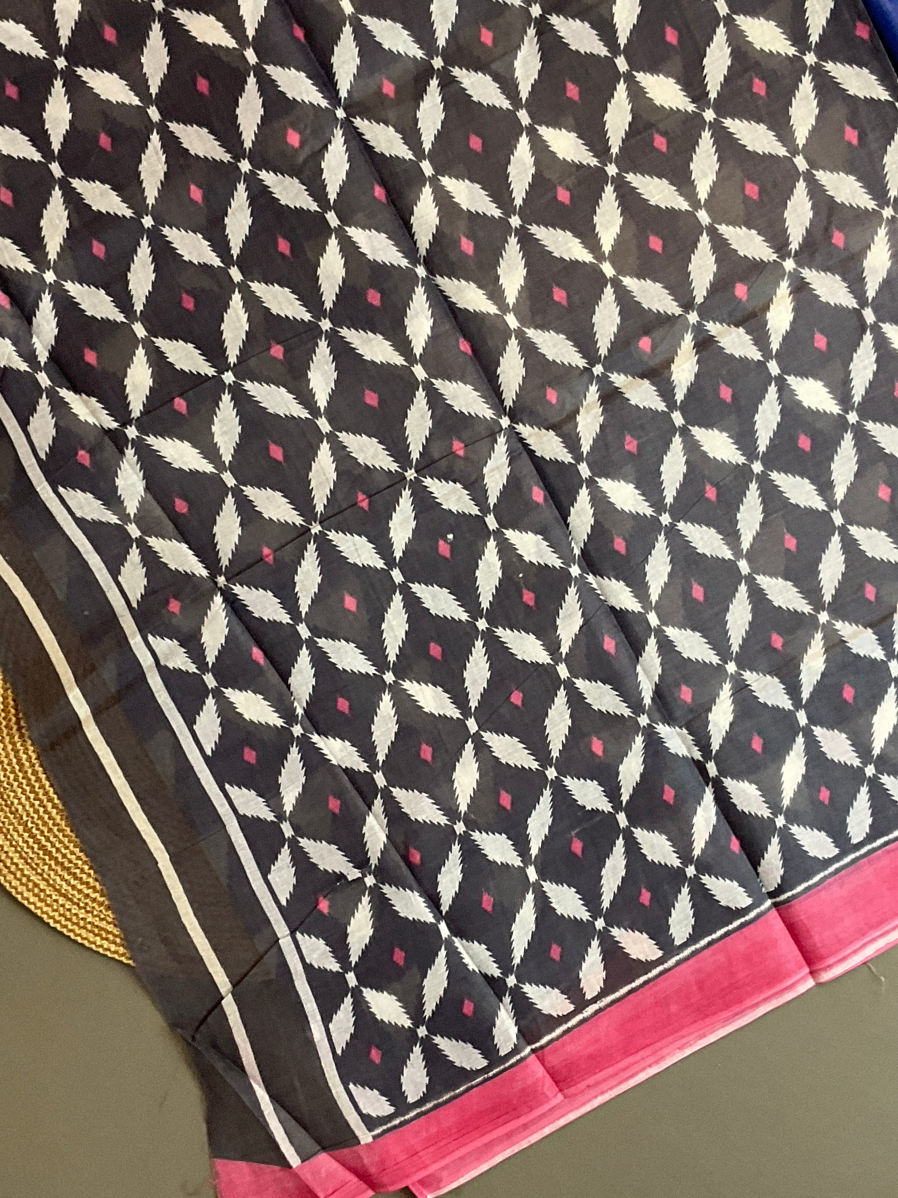 A close-up of a patterned fabric, likely a saree, featuring a dark charcoal gray base with a cream-colored ikat diamond motif, small pink diamond accents, and a vibrant pink border.