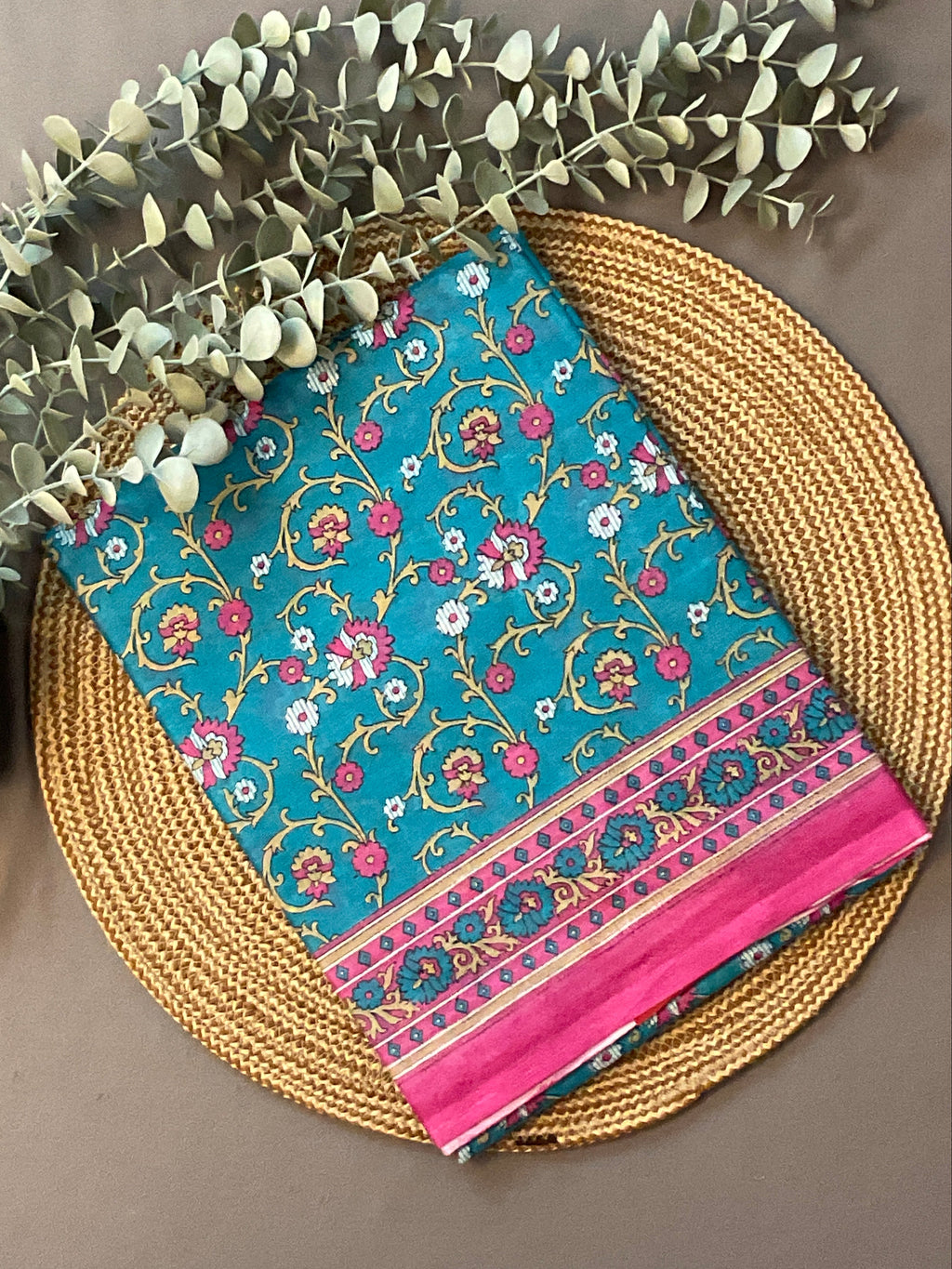 A folded teal blue fabric piece with a pink and gold floral vine pattern and a bright pink border, styled on a round woven straw mat with eucalyptus branches on a taupe background.