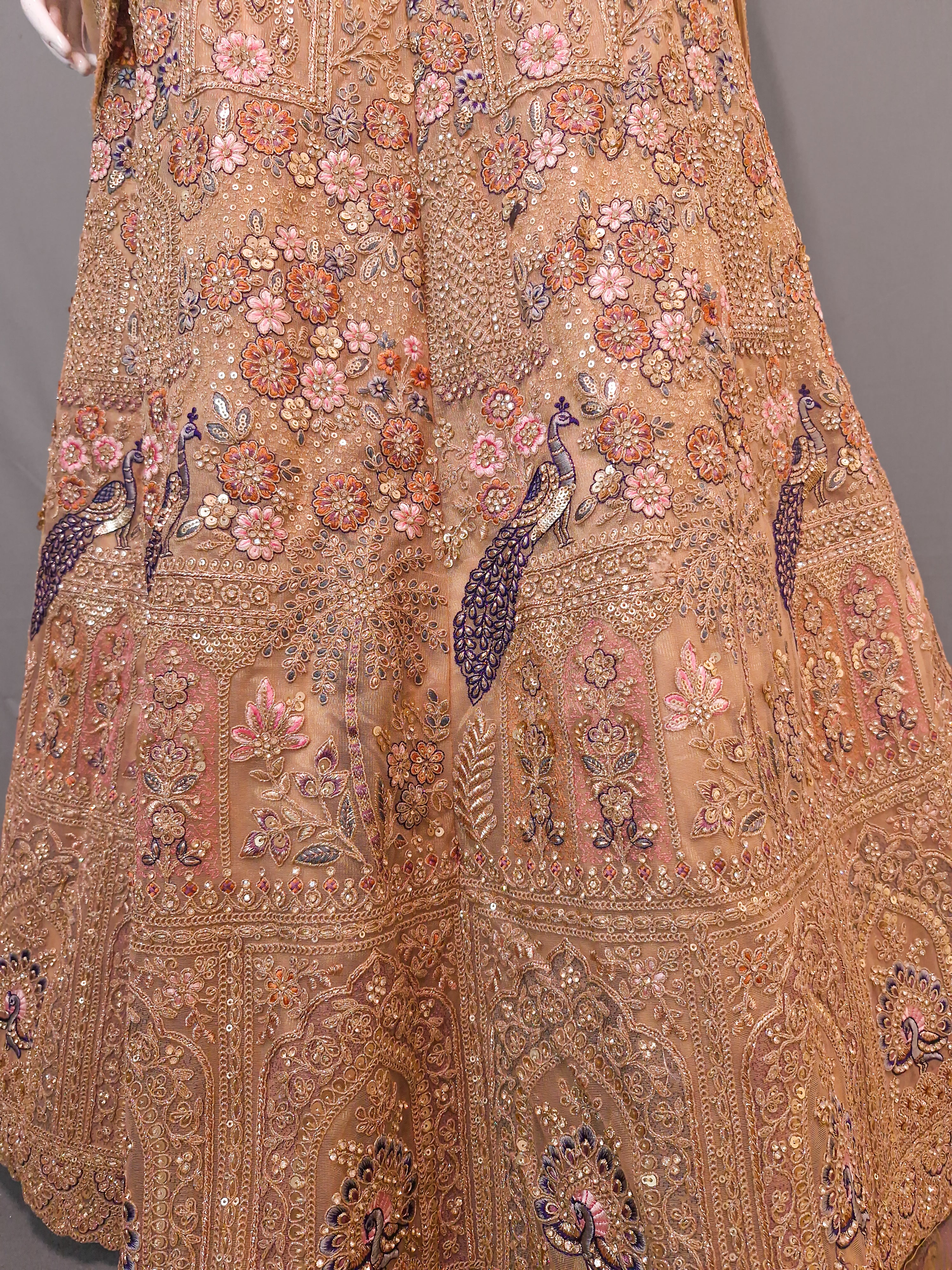 Close-up of an ornate embroidered traditional garment, likely a lehenga or skirt, in warm beige and peach tones with intricate floral, paisley, and peacock motifs in pink, purple, and metallic gold.