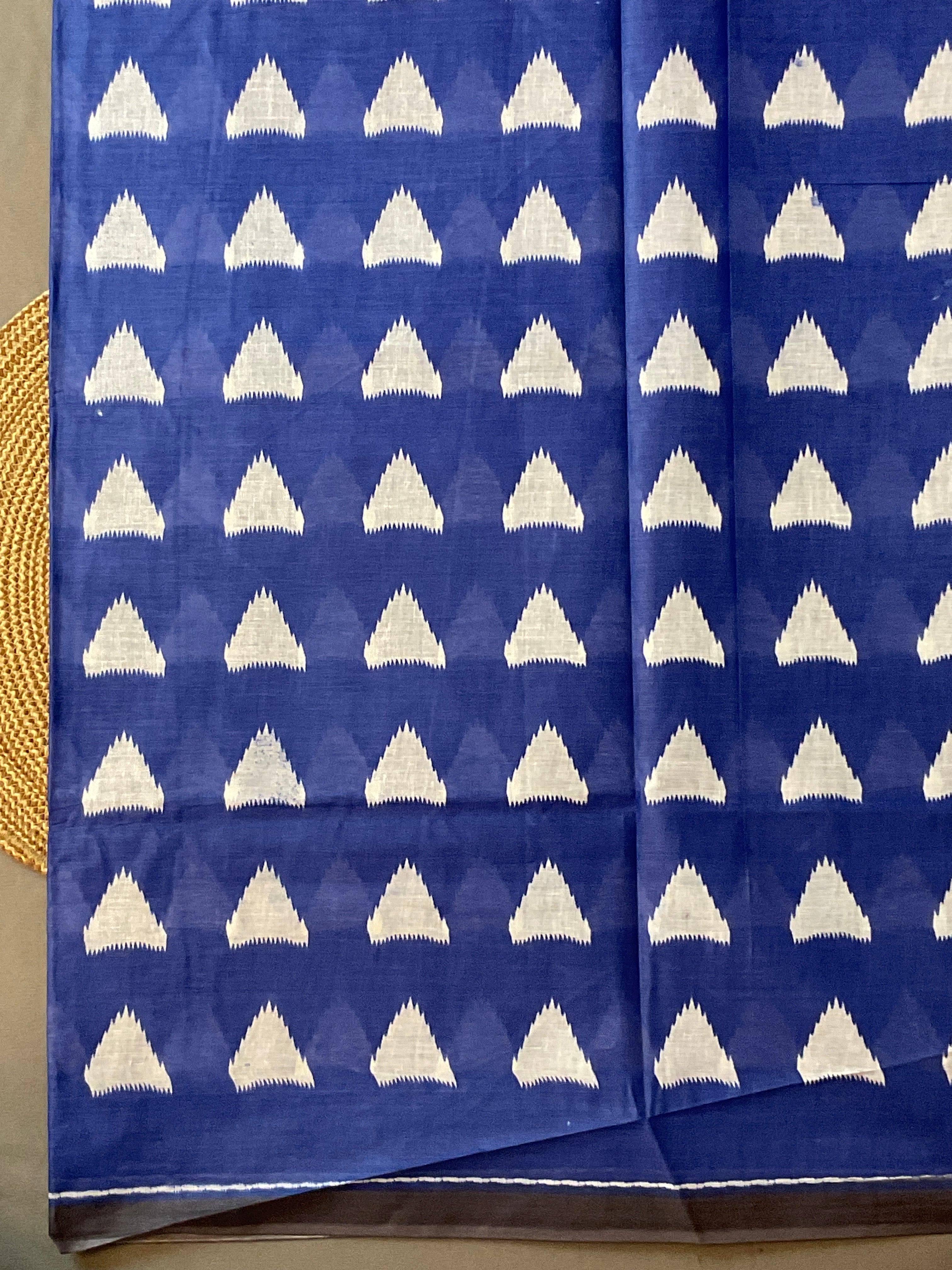 A folded royal blue fabric with a repeating white ikat-style triangle pattern, displayed flat with a small portion of a woven mat visible on the left edge.