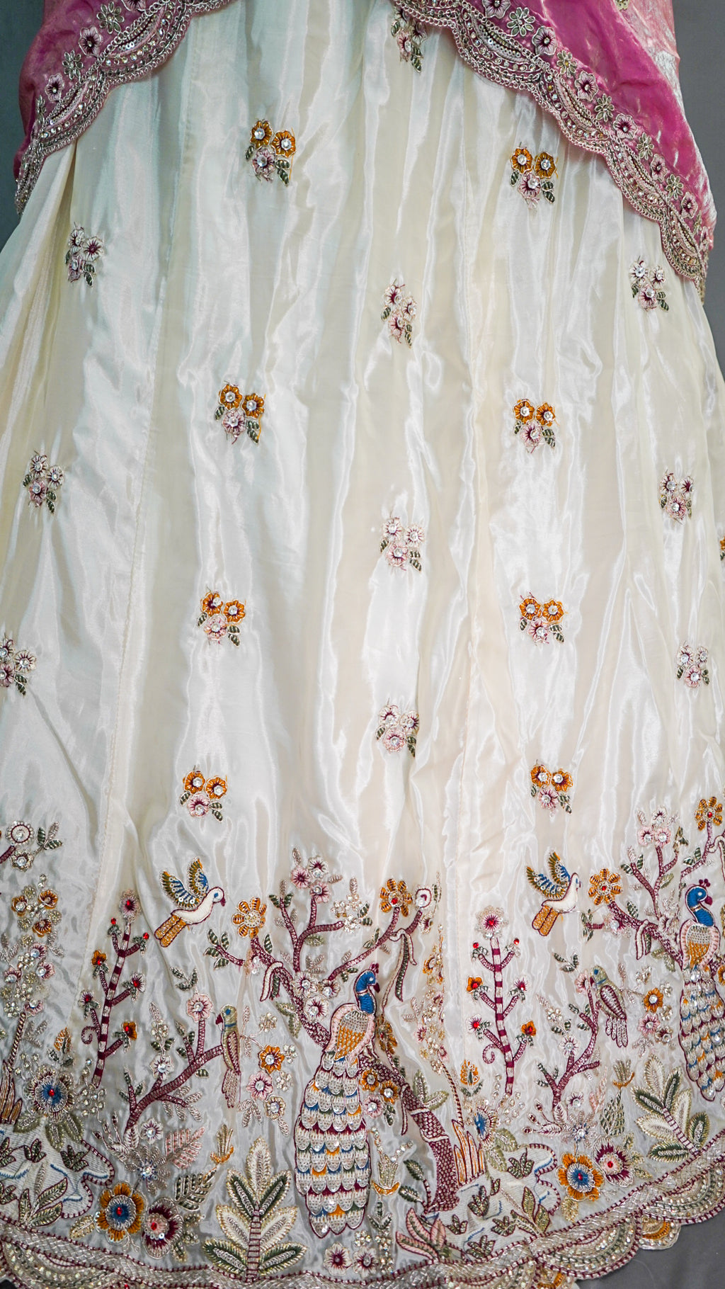An off-white silk saree body with intricate multicolor embroidery, featuring floral motifs and peacock designs near the border, with a pink border and a glimpse of a magenta dupatta.