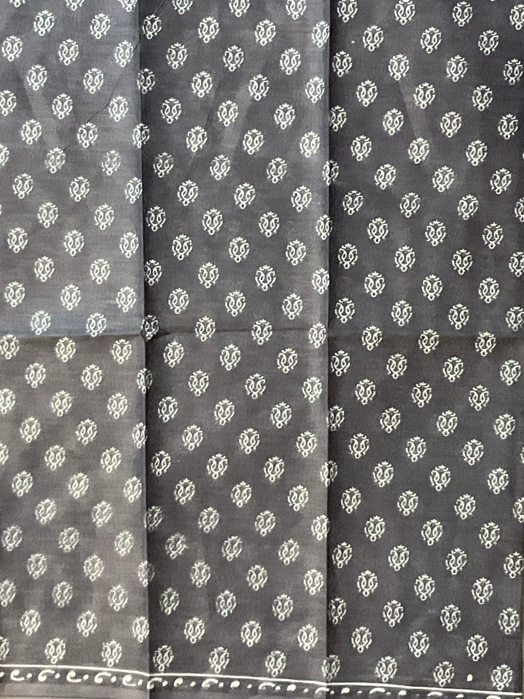 A dark gray fabric, likely a saree, featuring a repeating white block-printed floral pattern and a decorative border along the bottom edge.