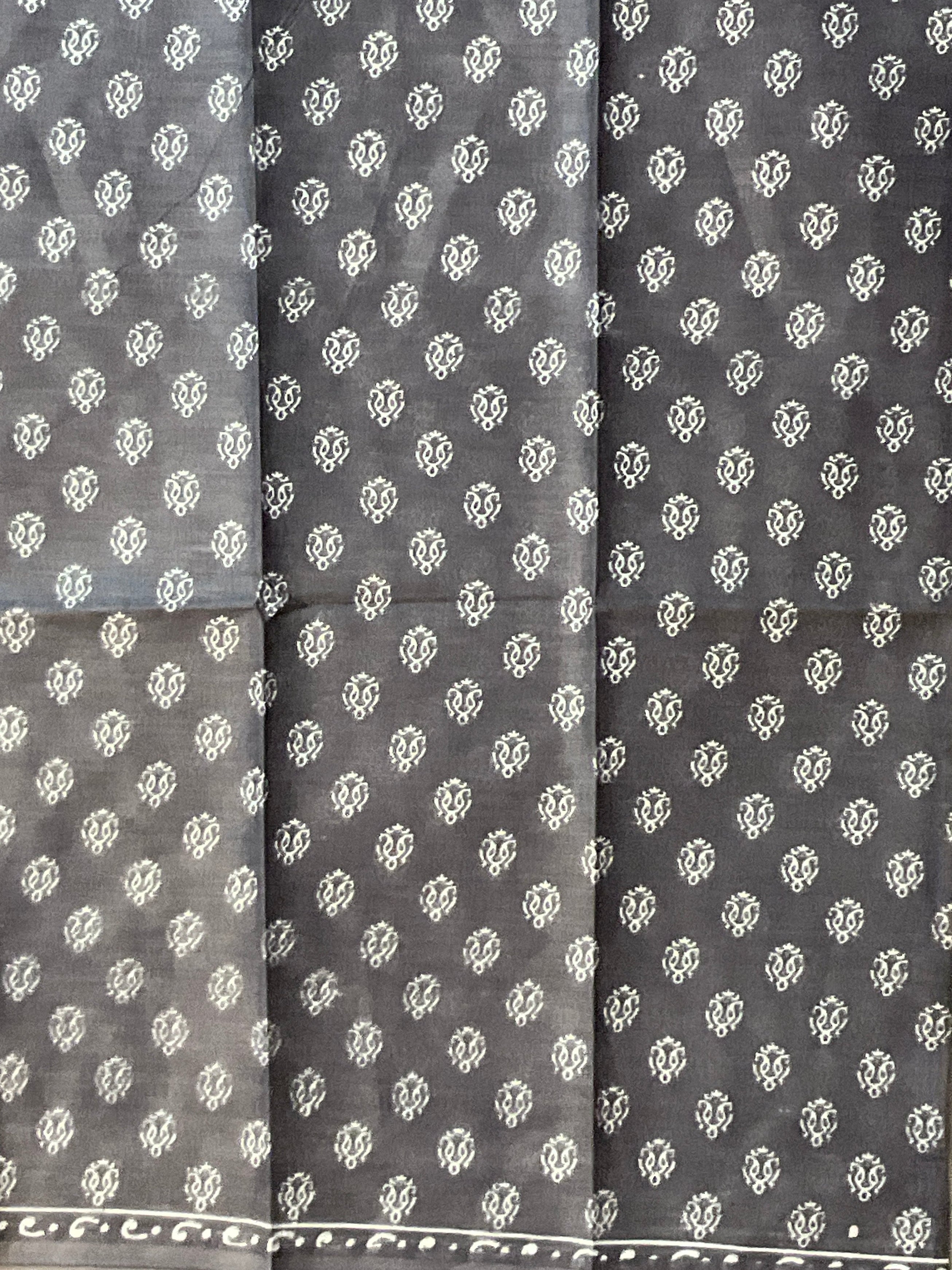 A dark gray fabric, likely a saree, featuring a repeating white block-printed floral pattern and a decorative border along the bottom edge.