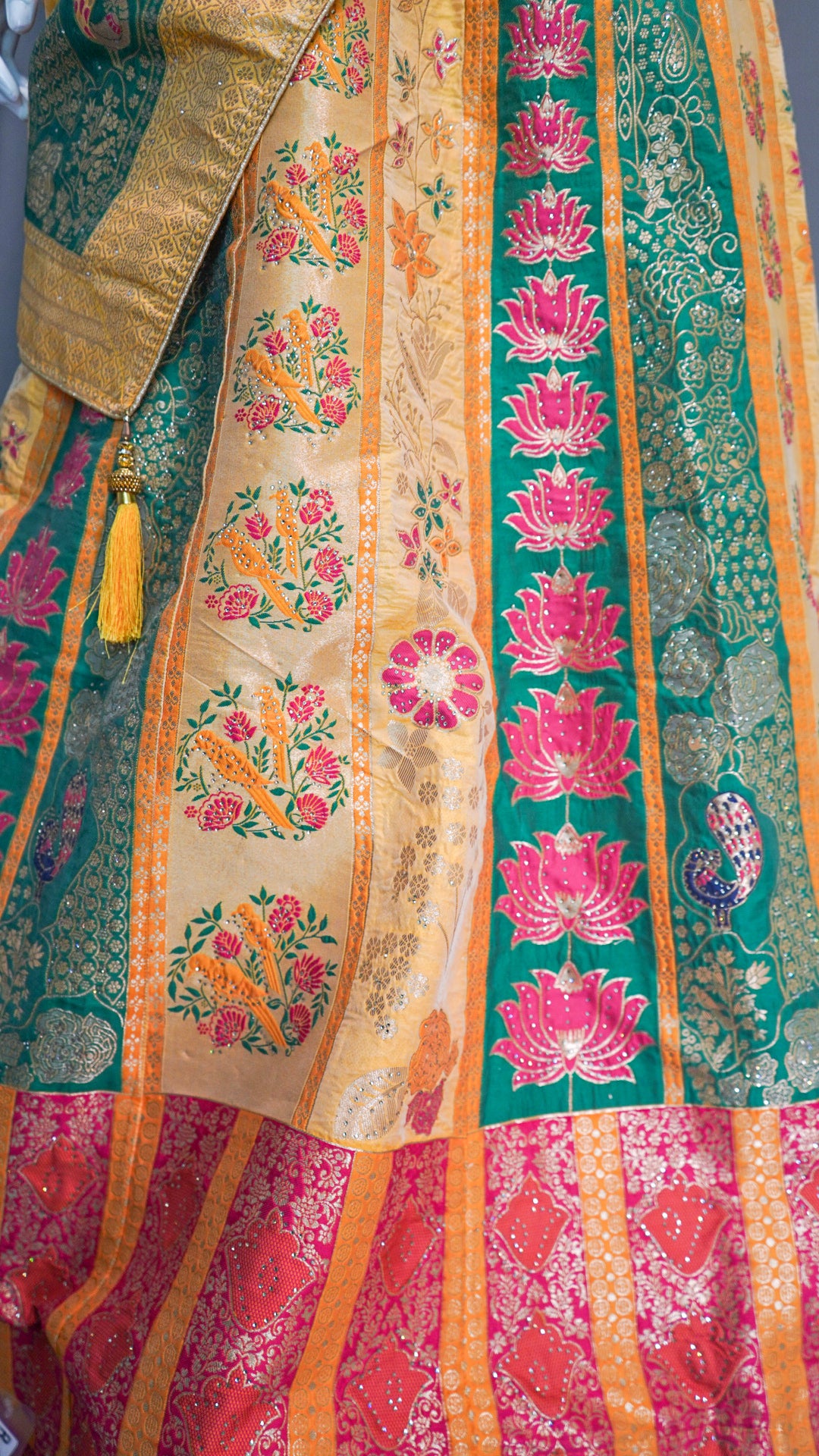 Close-up of a heavily embroidered traditional garment, likely a lehenga or sari, featuring intricate gold zari work on a beige base alongside vibrant green and pink floral panels with a yellow tassel.
